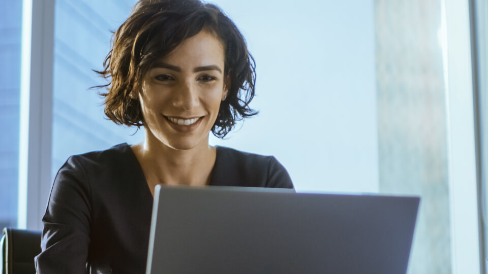 A woman sitting at an office desk watching a webinar on her laptop